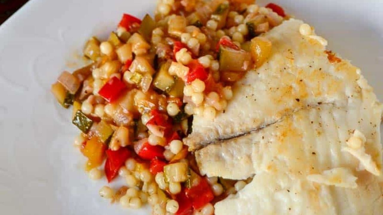 A cooked white fish fillet served next to a vegetable and pearl couscous medley on a white plate.