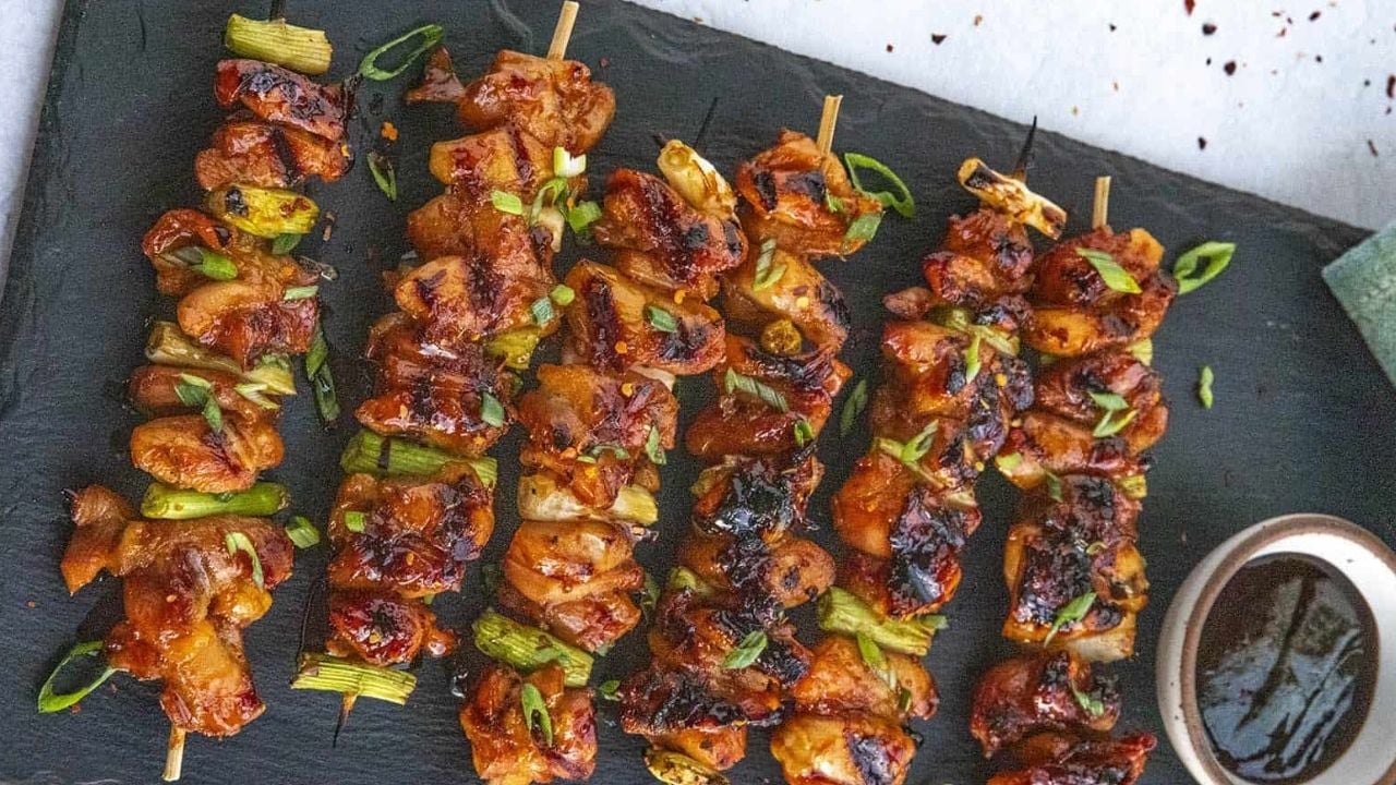 Grilled chicken skewers with vegetables are arranged on a dark serving tray, garnished with chopped green onions. A small bowl of dipping sauce sits beside them.