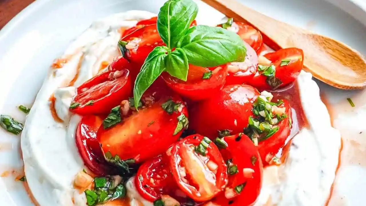 Fresh cherry tomatoes with basil and herbs on a bed of creamy white cheese, served on a white plate with a wooden spoon.