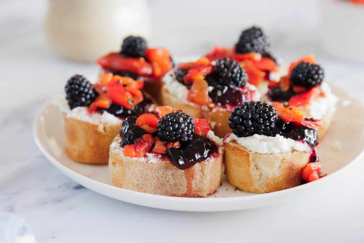 A plate of baguette slices topped with ricotta cheese, blackberry jam, diced red fruit, and fresh blackberries.