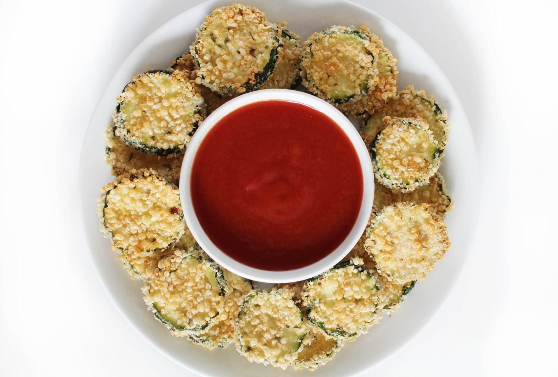 A plate of breaded zucchini chips arranged around a bowl of red dipping sauce.