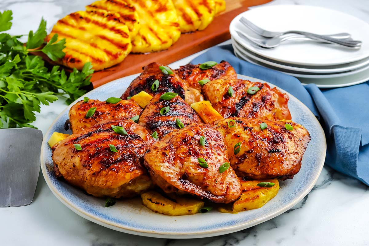 Grilled chicken thighs garnished with chopped green onions, served on a plate with grilled pineapple slices, with extra pineapple, fresh parsley, and stacked plates in the background.