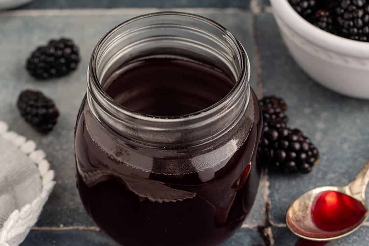 A glass jar filled with dark blackberry syrup sits on a table next to fresh blackberries, a silver spoon, and a white towel.