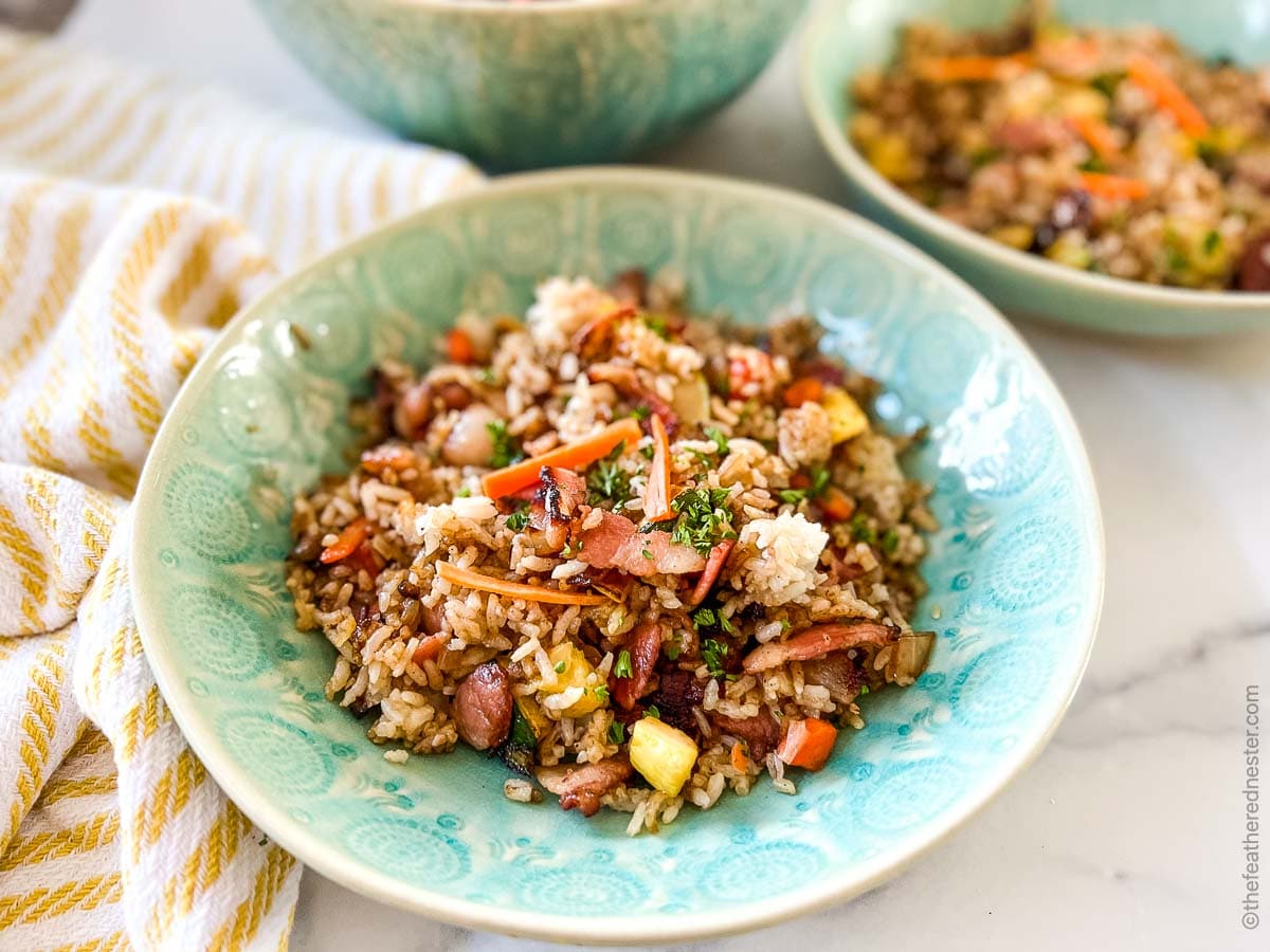A bowl of fried rice with vegetables, bacon, and scrambled eggs, served in a blue patterned dish on a marble surface.