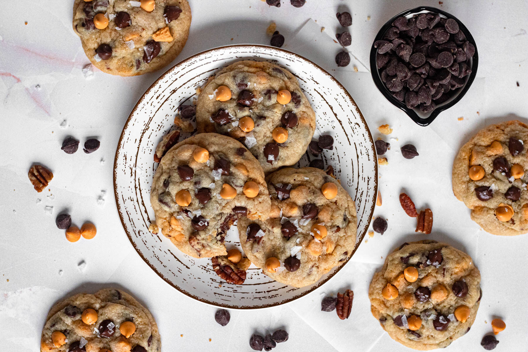 A plate of chocolate chip and butterscotch chip cookies is surrounded by more cookies, chocolate chips, and pecans on a white surface.