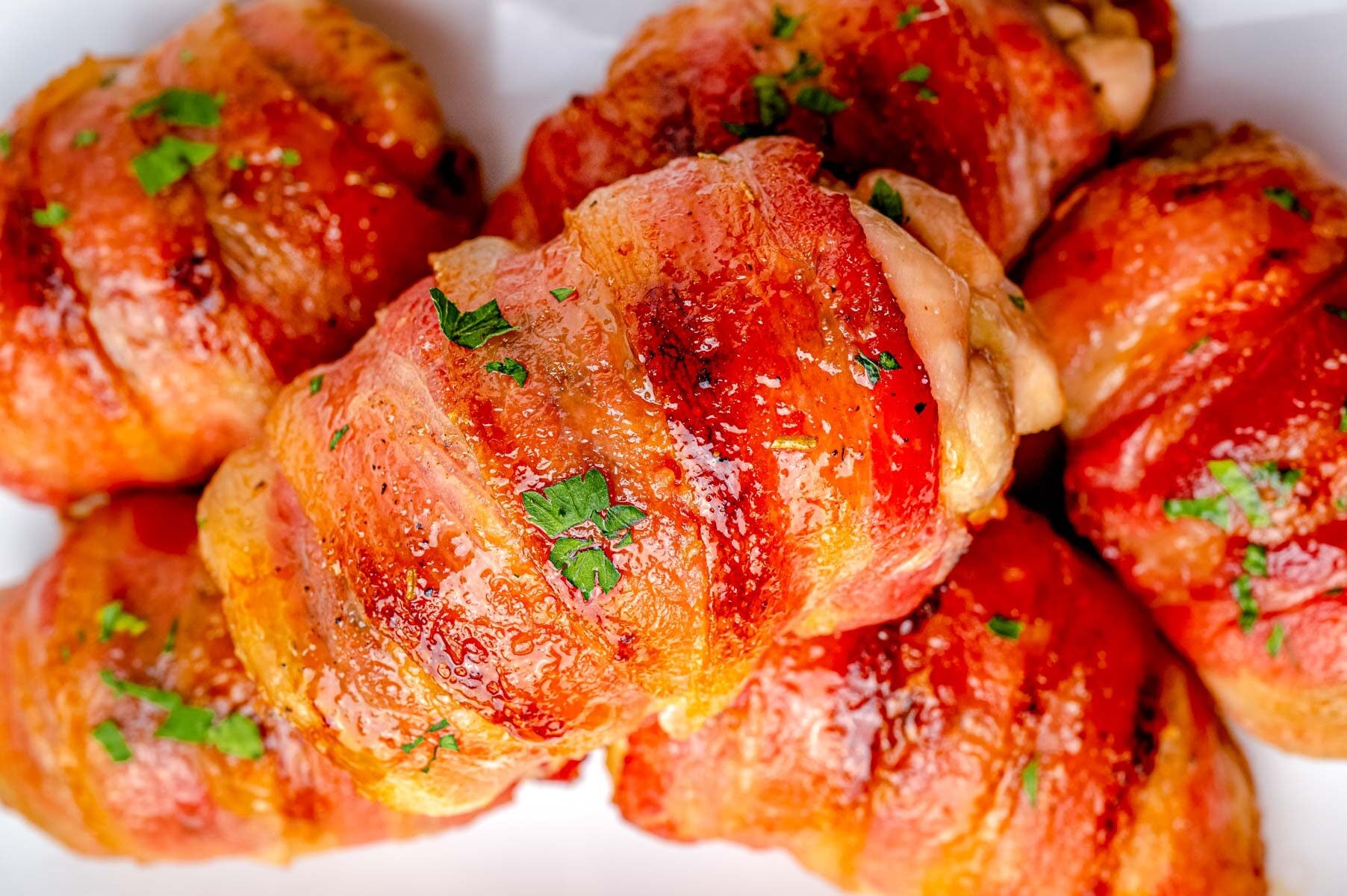 Close-up of several bacon-wrapped chicken pieces, garnished with chopped parsley, arranged on a white plate.