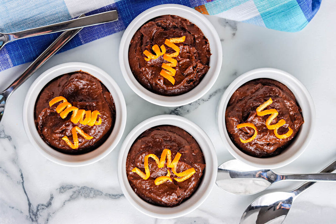Four small white ramekins filled with chocolate mousse are garnished with orange zest strips. Spoons and a blue napkin are placed nearby on a marble surface.