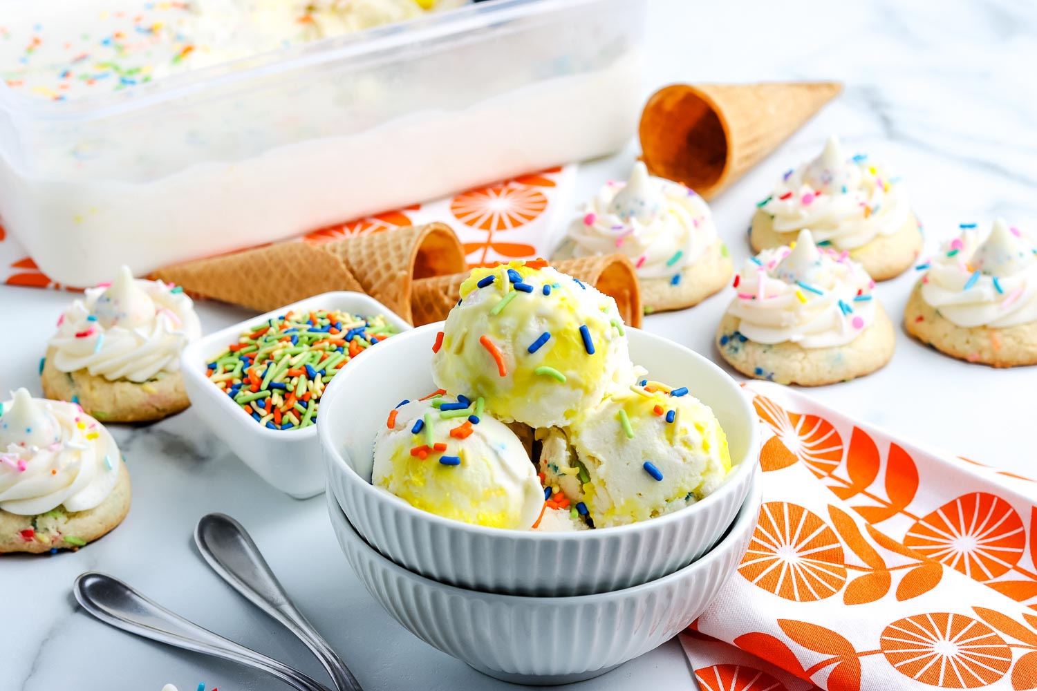 Two bowls of vanilla ice cream with rainbow sprinkles, surrounded by frosted cookies, sugar cones, a container of ice cream, sprinkles, and spoons on a white surface.