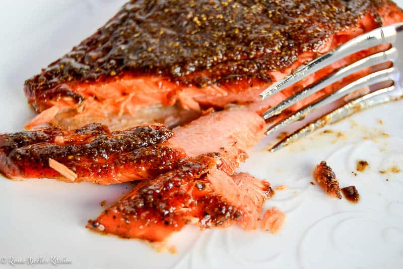 A close-up of a fork flaking a spicy, blackened piece of cooked salmon on a white plate.