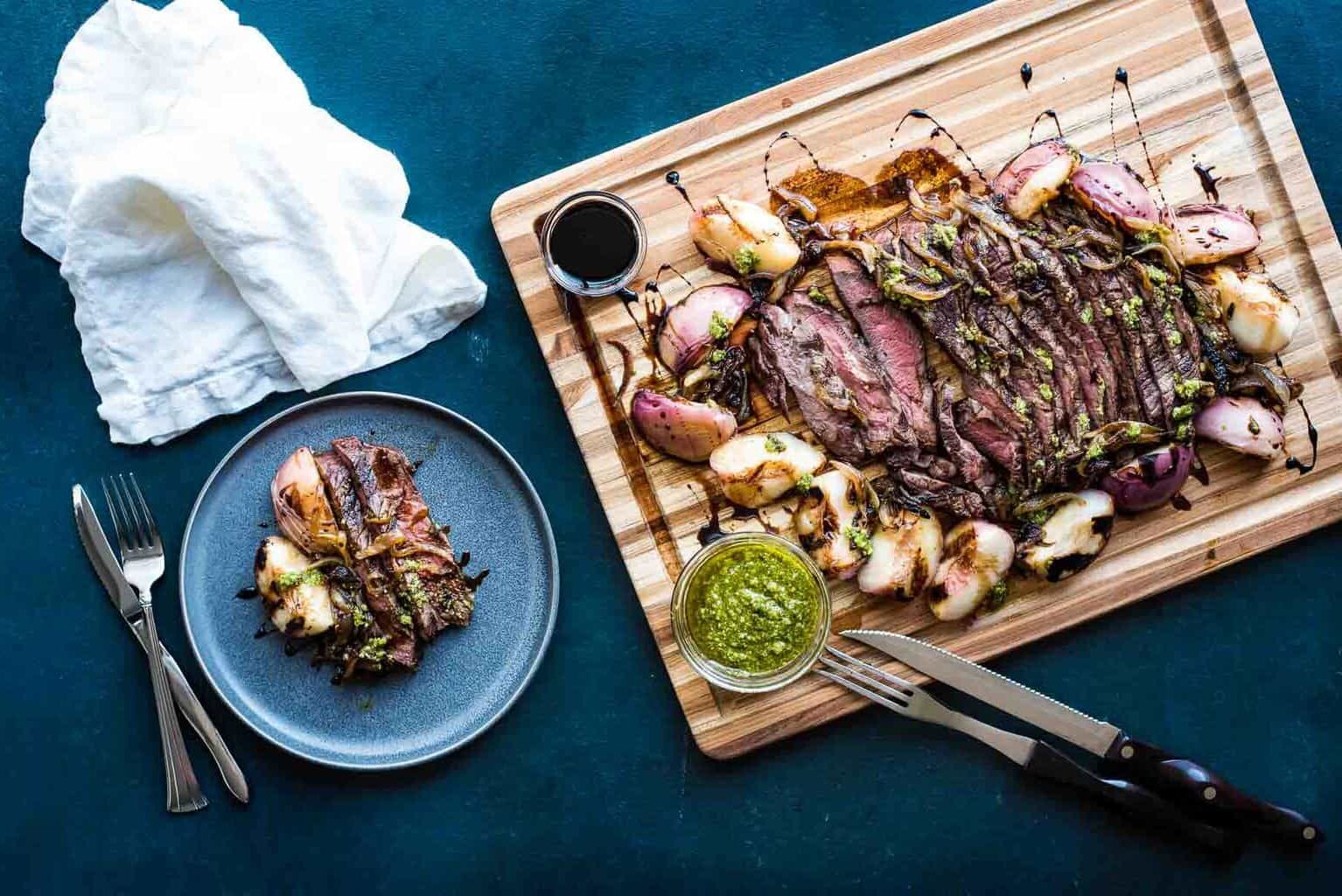 A wooden board with sliced steak, grilled onions, sauce, and chimichurri sits beside a plate with a portion of the same meal; utensils and a napkin are nearby.