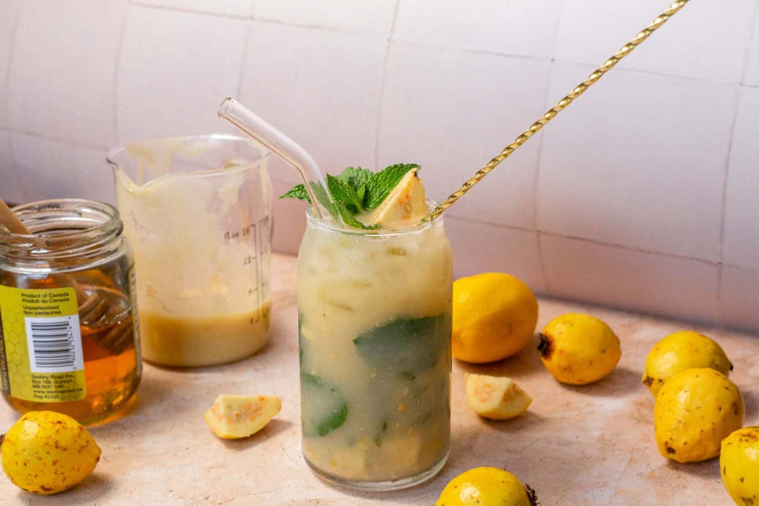 A glass of yellow guava drink with ice, mint, and a glass straw sits on a table surrounded by fresh guavas, honey, and a small pitcher.
