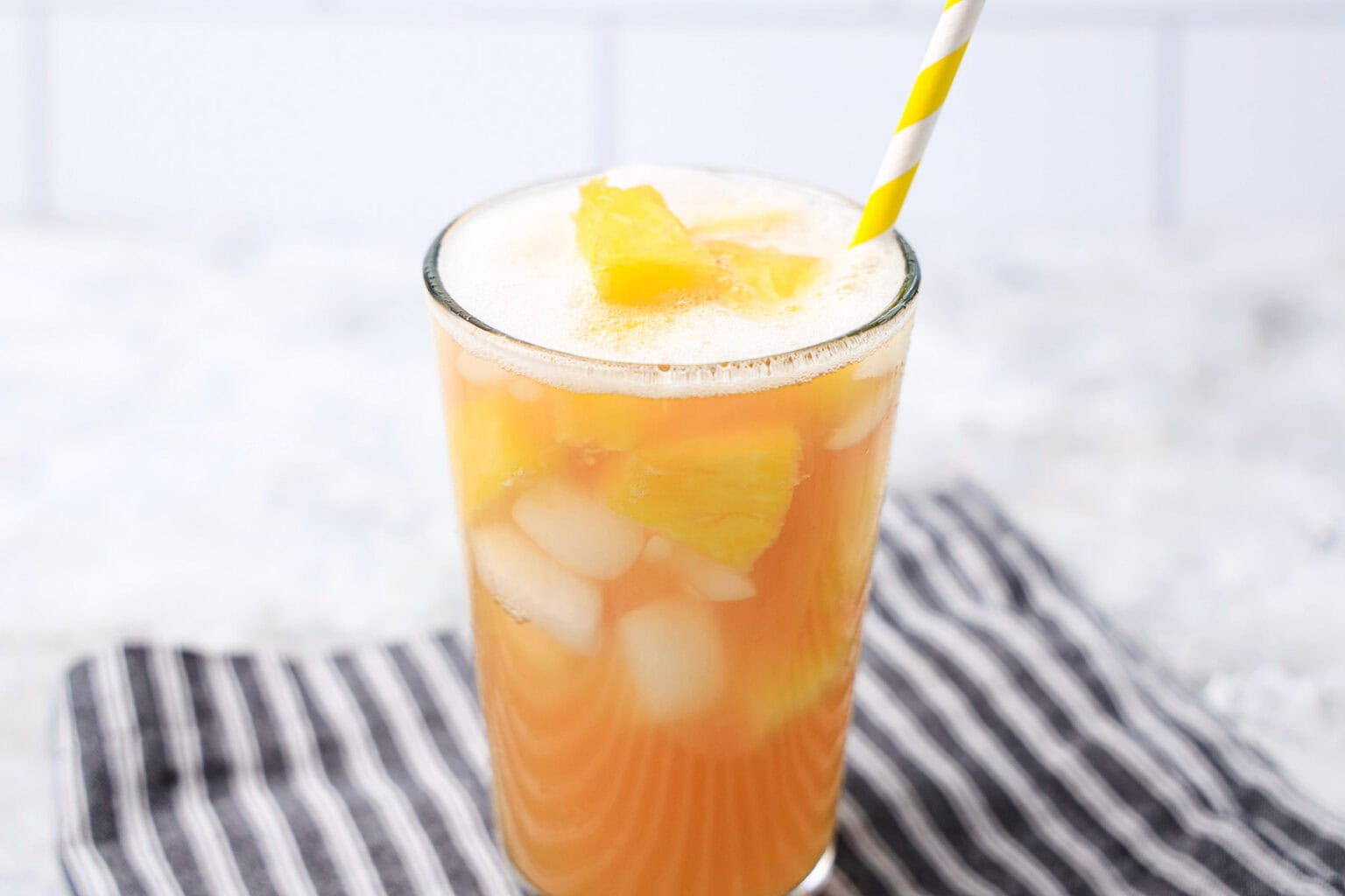 A glass of iced pineapple juice with pineapple chunks and a yellow-striped straw, placed on a black and white striped cloth.
