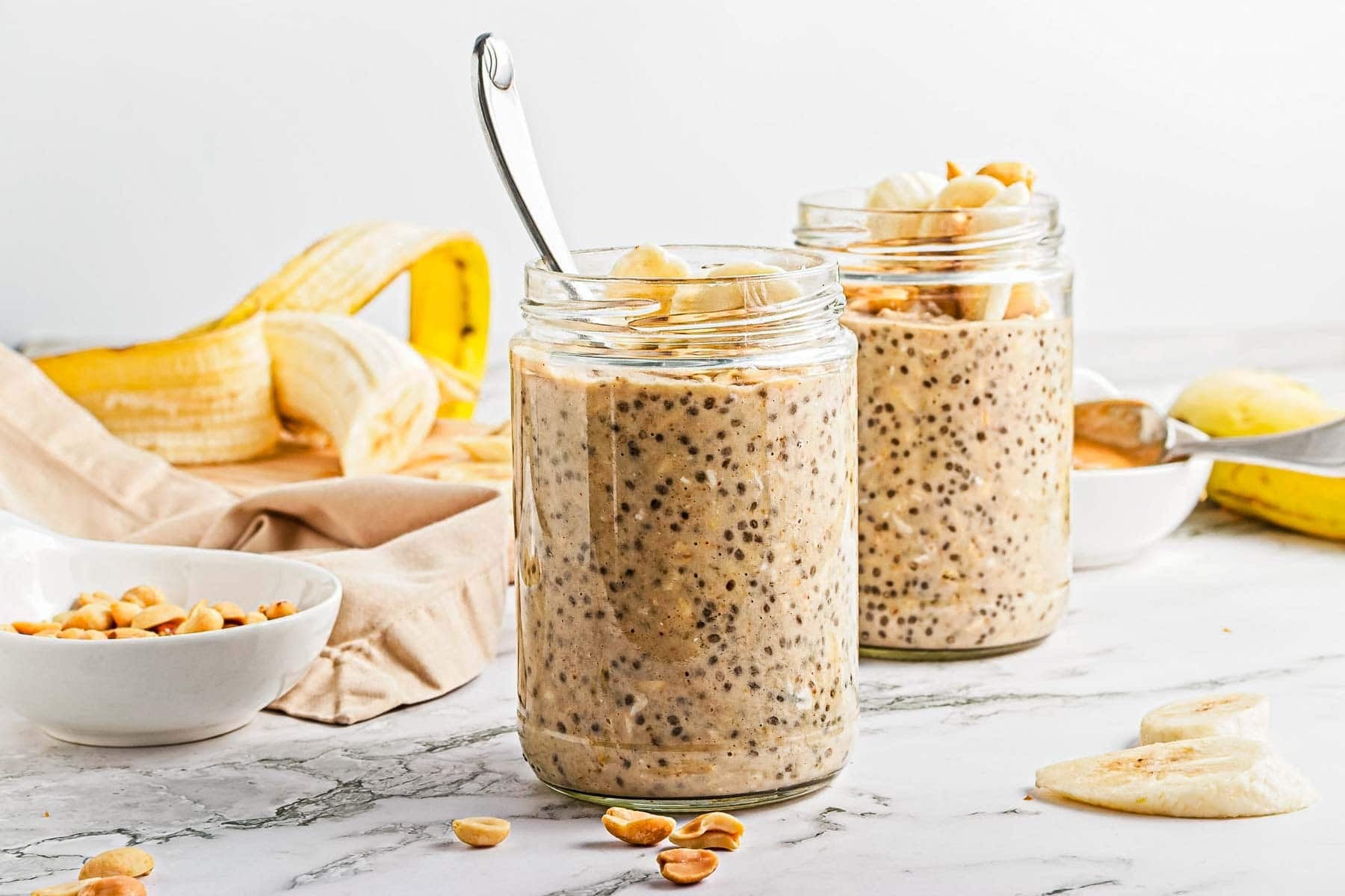 Two glass jars filled with overnight oats topped with banana slices and peanuts, with a spoon in one jar. Sliced bananas and a bowl of peanuts are in the background.