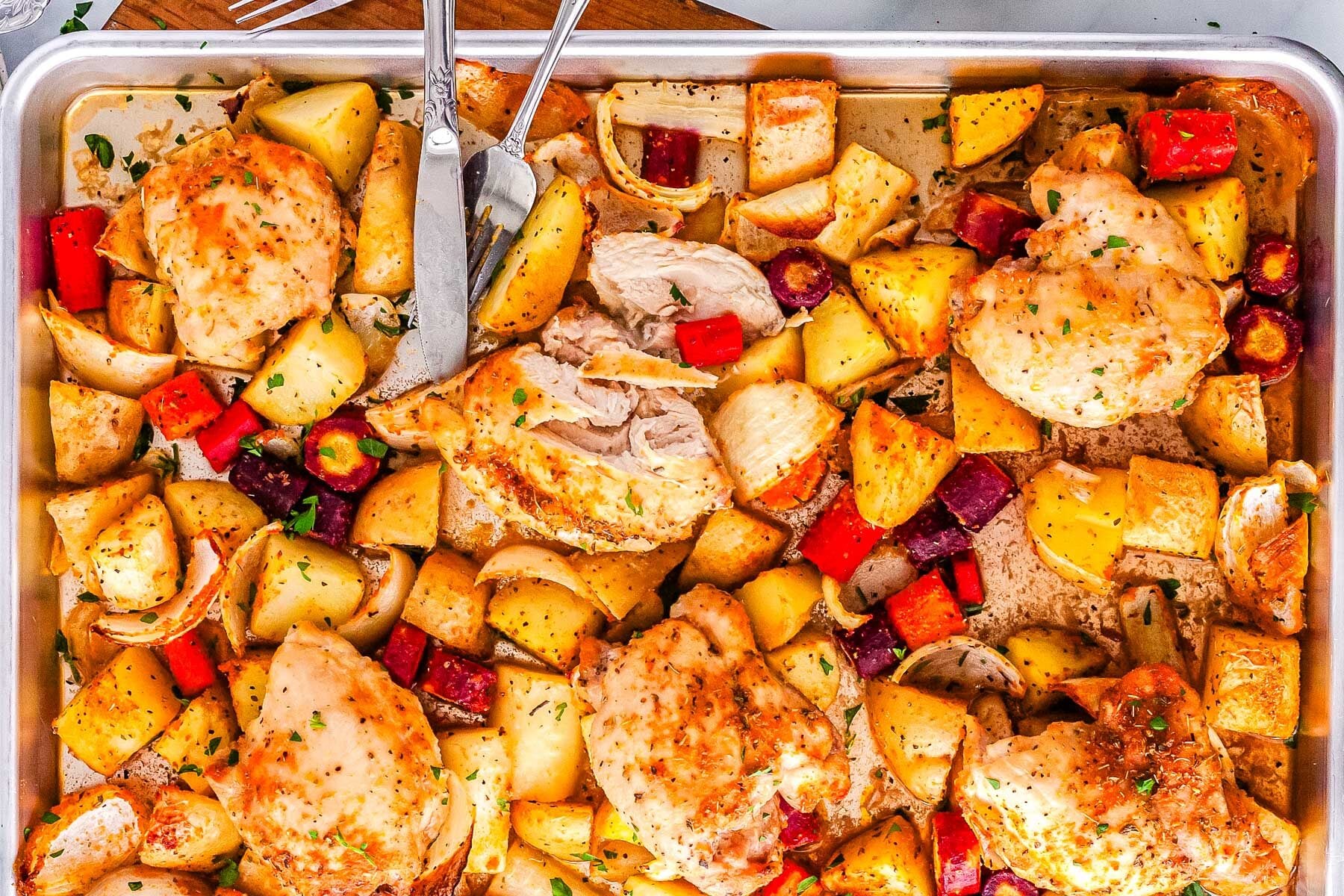 Sheet pan with baked chicken breasts, diced potatoes, carrots, and onions, seasoned and garnished with herbs. A fork and knife are on the tray, one chicken breast is sliced.