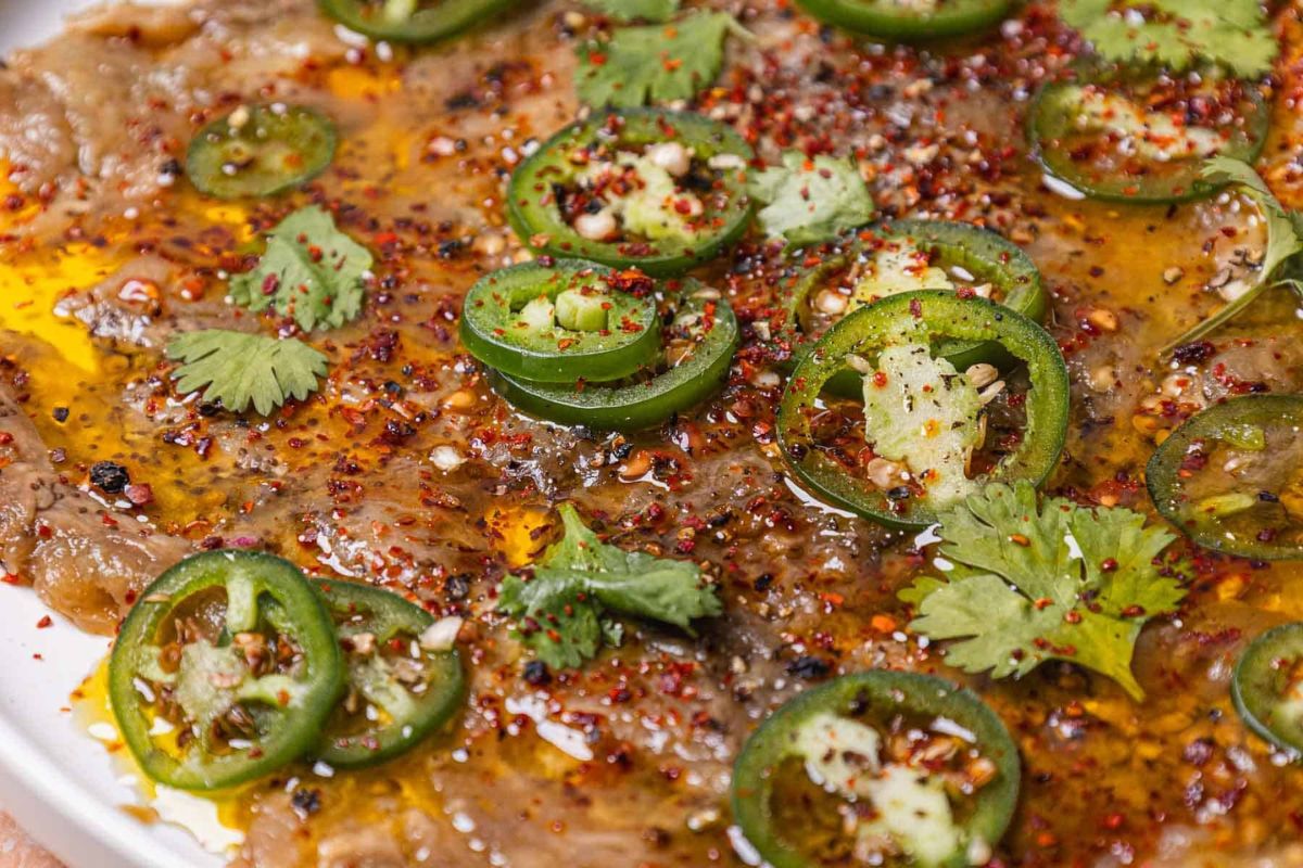 Thinly sliced raw beef topped with jalapeño slices, cilantro, chili flakes, cracked pepper, and drizzled with oil on a white plate.