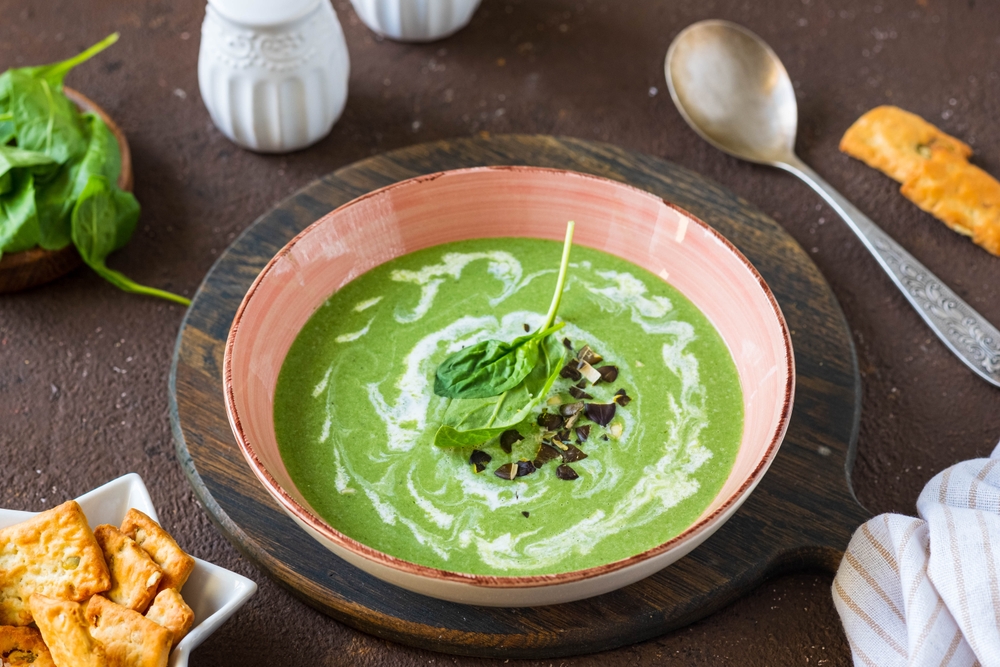 A bowl of creamy green spinach soup garnished with a spinach leaf and chopped herbs, served on a wooden tray with crackers and a spoon nearby.