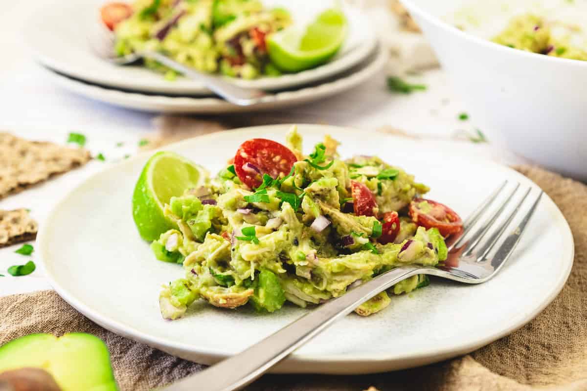 A plate of guacamole salad with chopped tomatoes, red onion, herbs, and a lime wedge, served with a fork on a white plate.