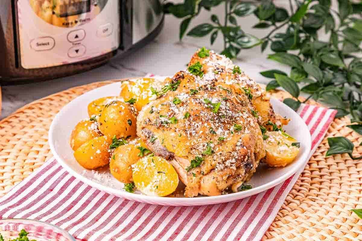 A plate of cooked chicken and yellow potatoes garnished with herbs and grated cheese, placed on a striped napkin next to a slow cooker.