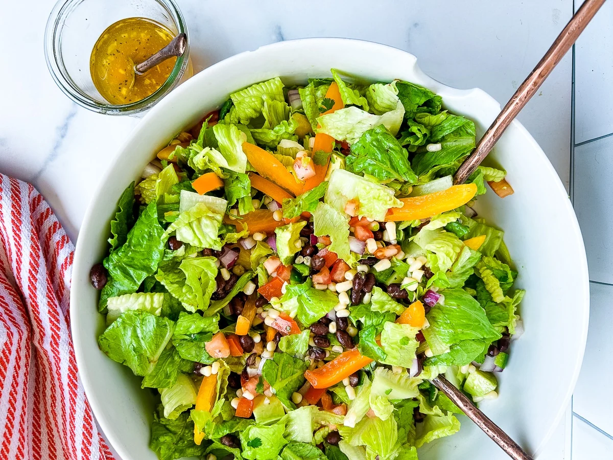 A bowl of chopped salad with lettuce, black beans, corn, diced tomatoes, and sliced yellow peppers, next to a jar of vinaigrette and a red-striped towel.