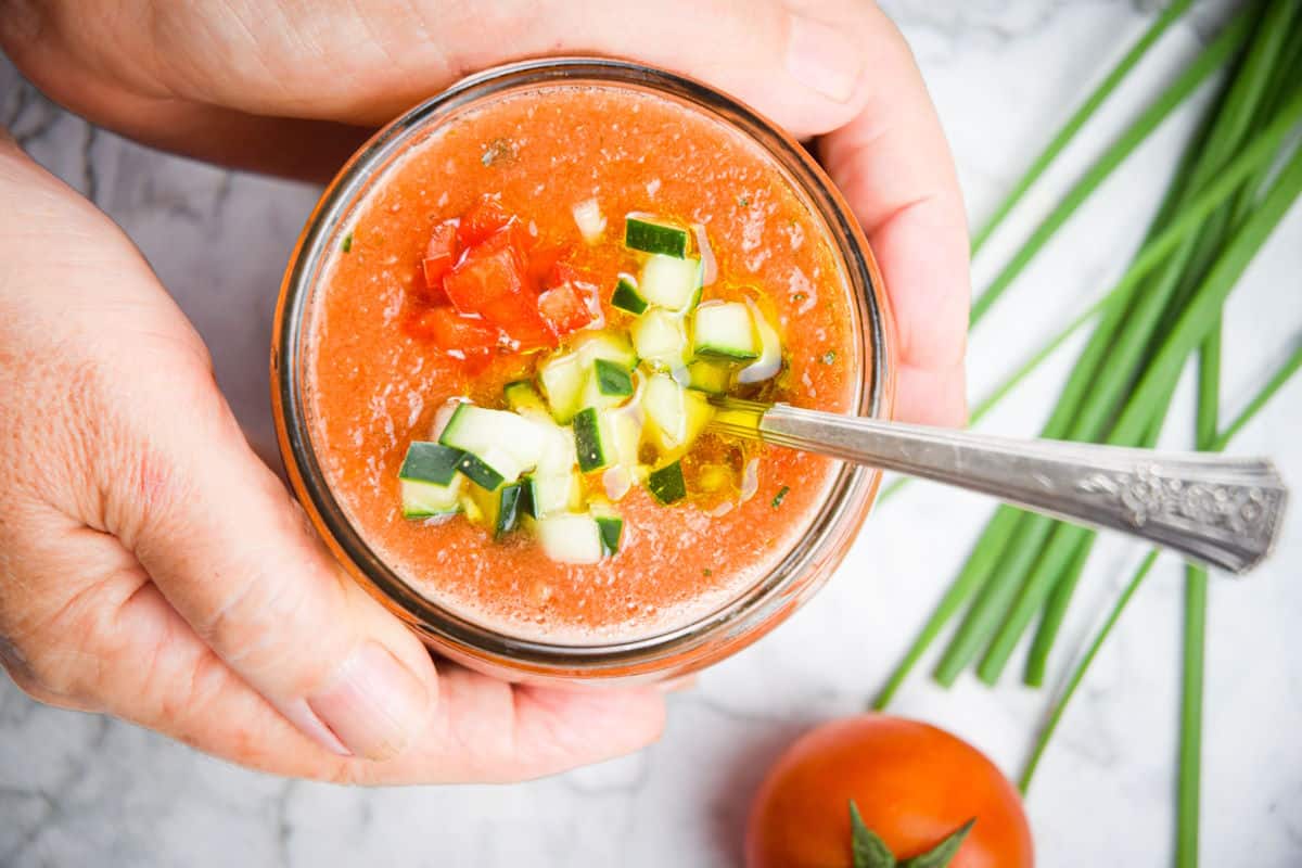 A person holds a bowl of gazpacho topped with diced cucumber and tomato, with a spoon inside. Chives and a tomato are on the white surface nearby.