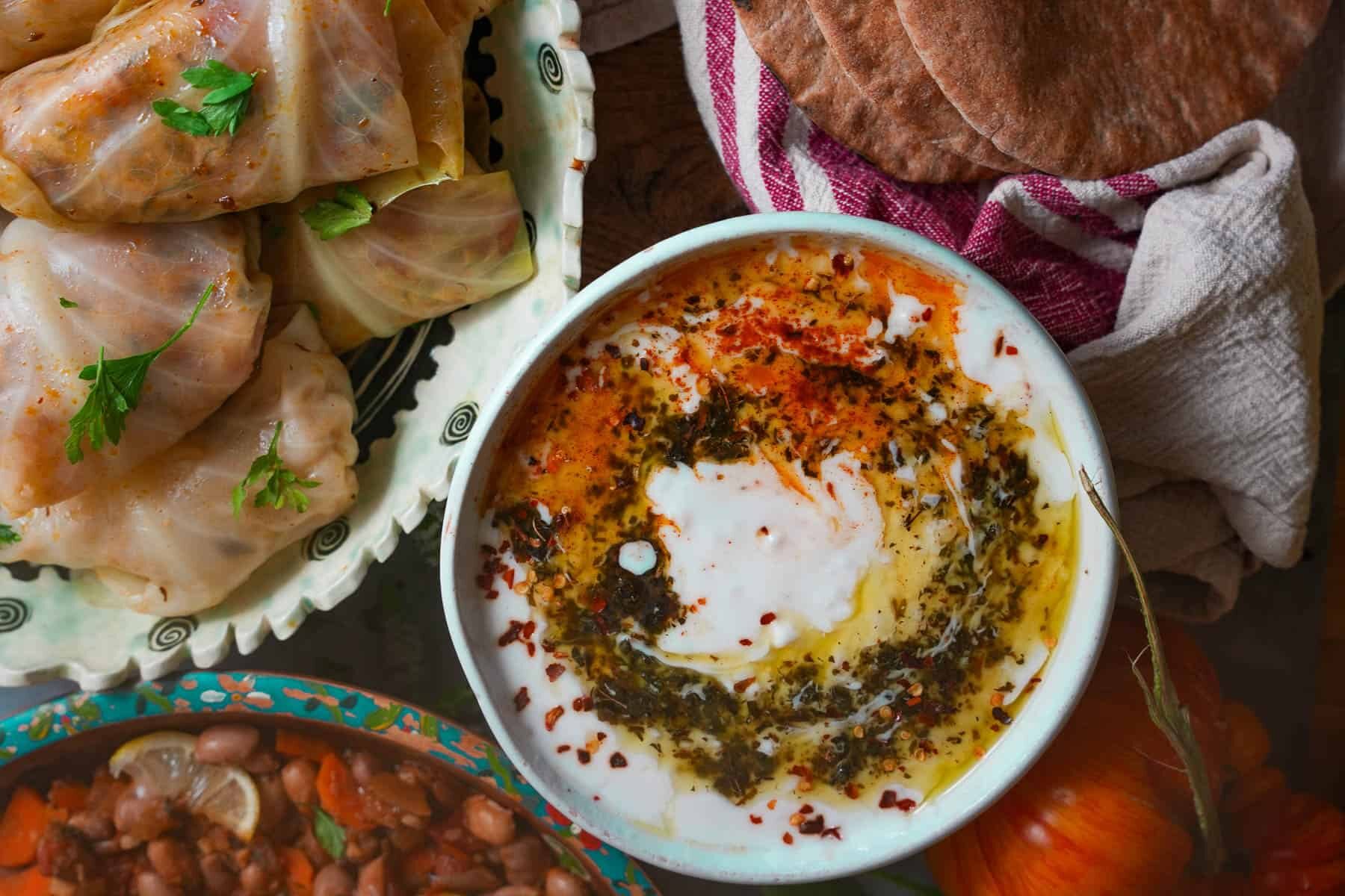 A bowl of yogurt dip with spices and oil, surrounded by stuffed cabbage rolls, pita bread, and a dish of beans.