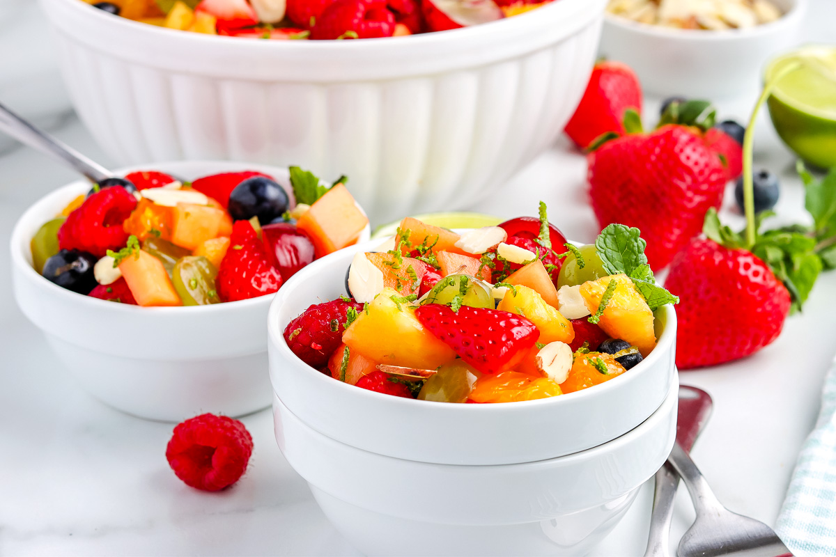 Bowls of fresh fruit salad with strawberries, raspberries, blueberries, peaches, sliced almonds, and mint leaves on a white surface, with whole strawberries and utensils nearby.