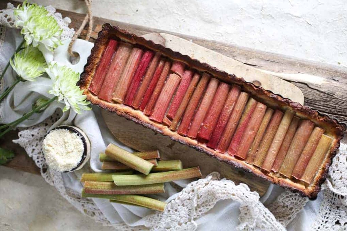 Rectangular rhubarb tart with neatly arranged rhubarb stalks, placed on a wooden board with lace cloth, fresh flowers, and a small bowl of powdered sugar nearby.