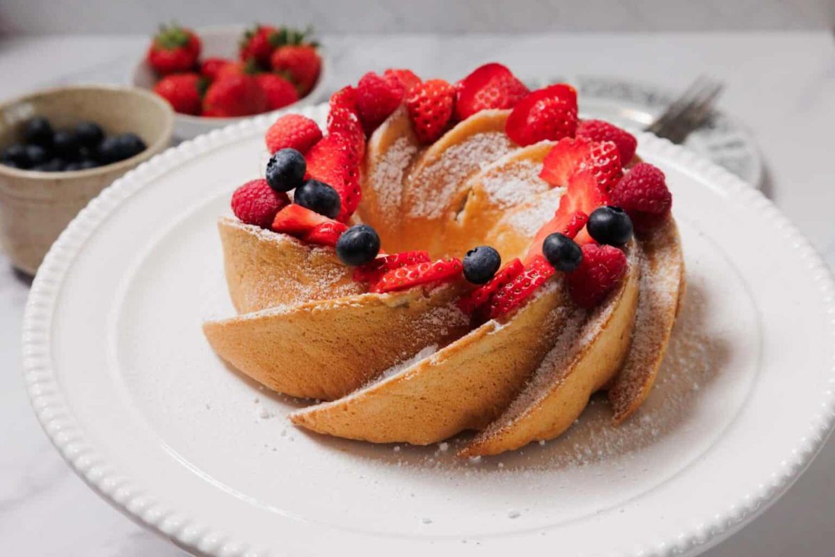 A bundt cake topped with powdered sugar, sliced strawberries, whole raspberries, and blueberries on a white plate. Bowls of berries are in the background.