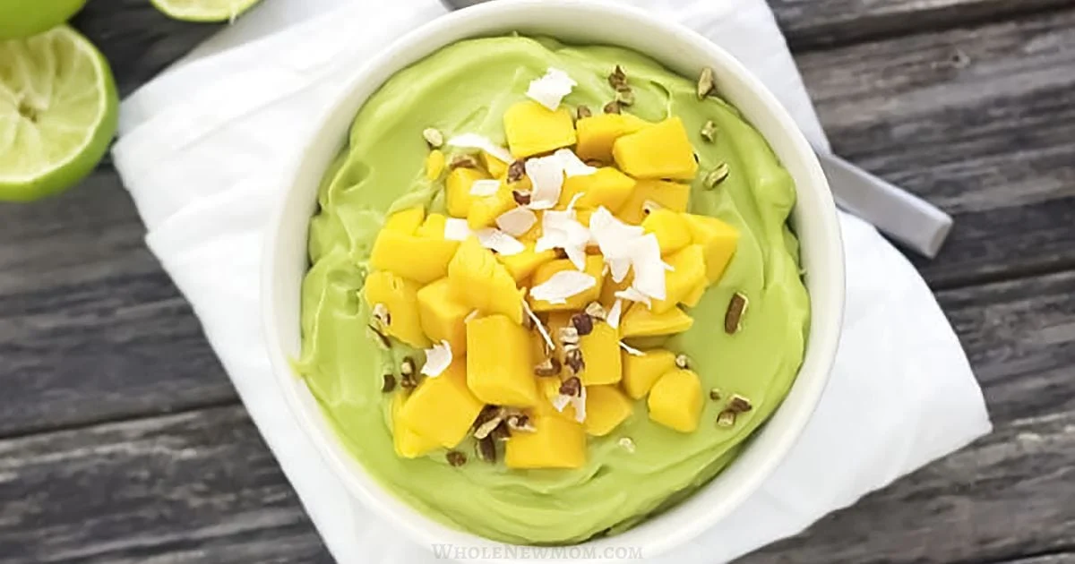 A white bowl filled with green avocado mousse, topped with diced mango, coconut flakes, and chopped nuts, on a white cloth and wooden surface.