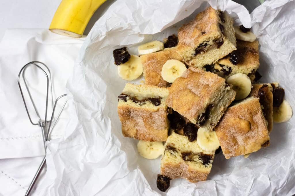 Squares of banana cake with pieces of dried fruit are arranged on white parchment paper, with banana slices and a banana in the background. An egg beater lies nearby.
