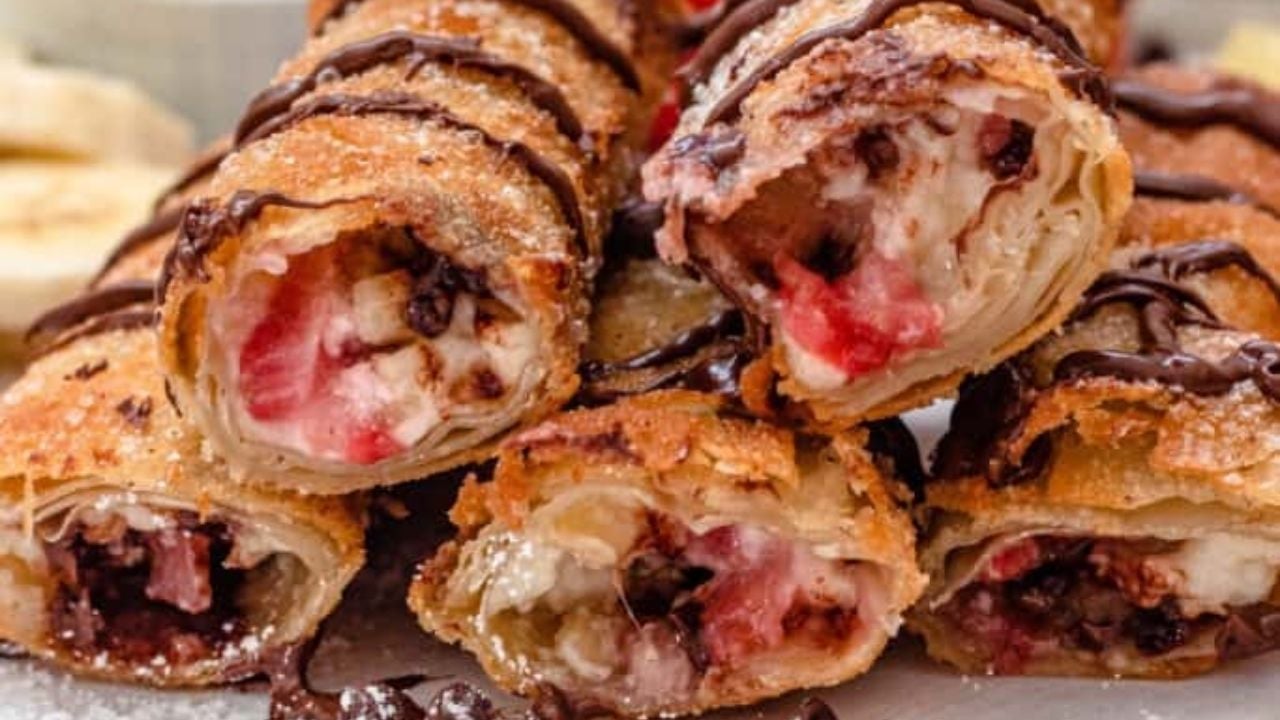 Close-up of stacked fried pastry rolls filled with strawberries, bananas, and chocolate, drizzled with chocolate sauce.