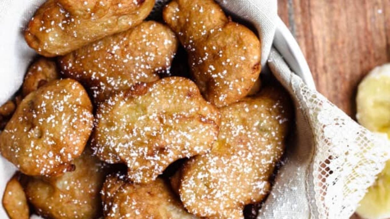 A basket lined with cloth filled with golden-brown, heart-shaped fritters dusted with powdered sugar, placed on a wooden surface.
