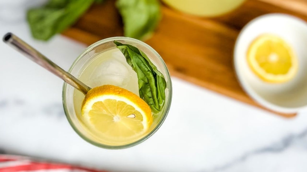 A glass of iced lemonade with a metal straw, garnished with a lemon slice and fresh basil leaf, sits on a white surface next to a bowl with a lemon slice and a wooden board.