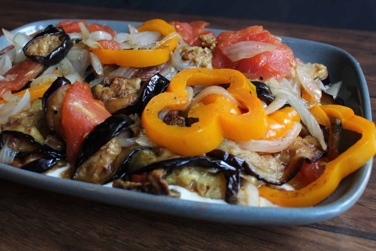 A rectangular dish filled with roasted vegetables including yellow bell pepper rings, tomato slices, onions, and eggplant on a wooden surface.