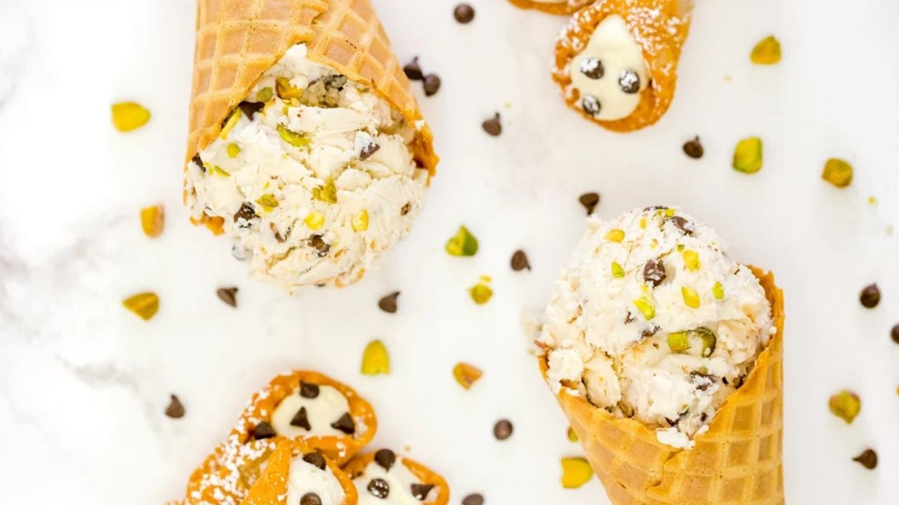 Waffle cones filled with cannoli-inspired ice cream, topped with chocolate chips and chopped pistachios, with scattered chocolate chips and pistachio pieces on a white surface.