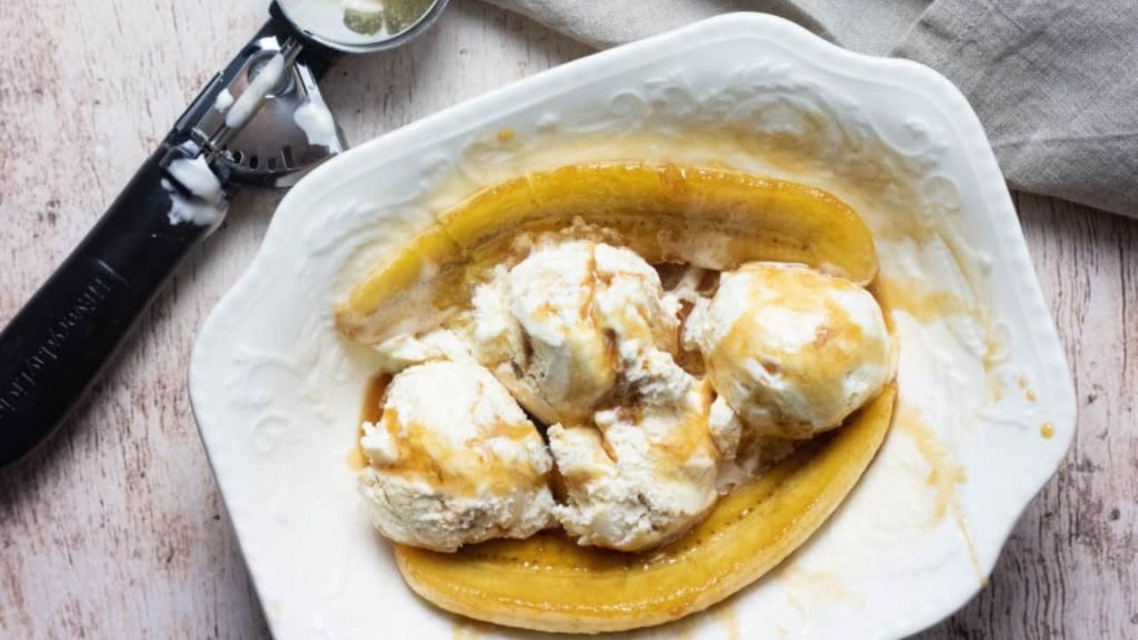 A banana split dessert with three scoops of vanilla ice cream, caramel sauce, and a halved banana on a white plate next to an ice cream scoop.