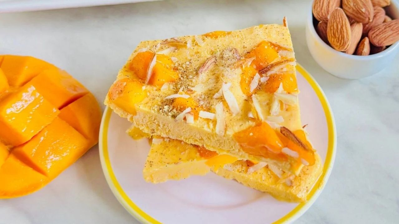Two pieces of mango and almond dessert are stacked on a plate, with diced mango and a bowl of whole almonds beside them on a white surface.