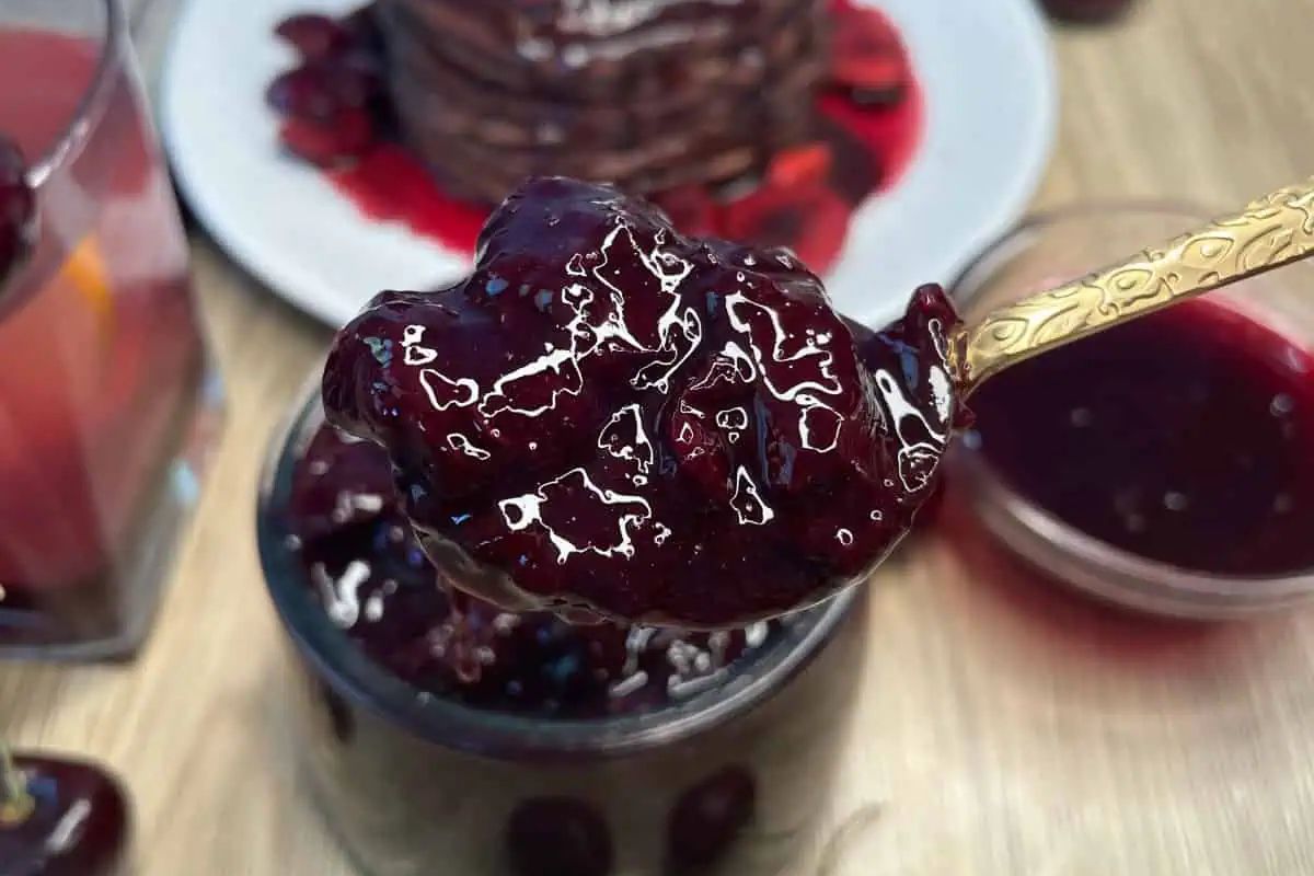 A spoonful of thick, glossy berry jam is held above a jar, with pancakes topped with the same jam visible in the background.