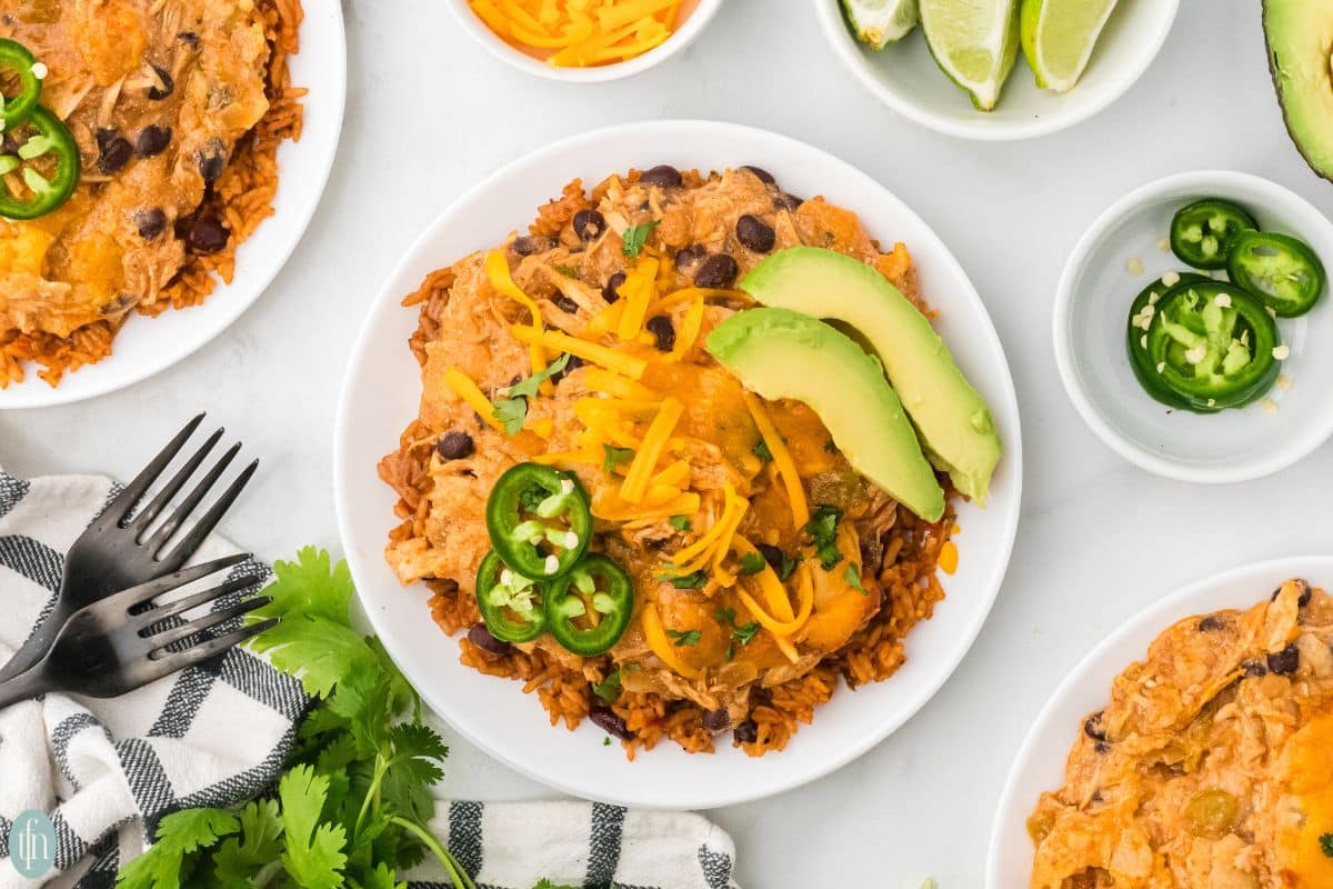 A plate of Mexican rice casserole topped with shredded cheese, sliced avocado, and jalapeño slices, surrounded by lime wedges, cilantro, and utensils.
