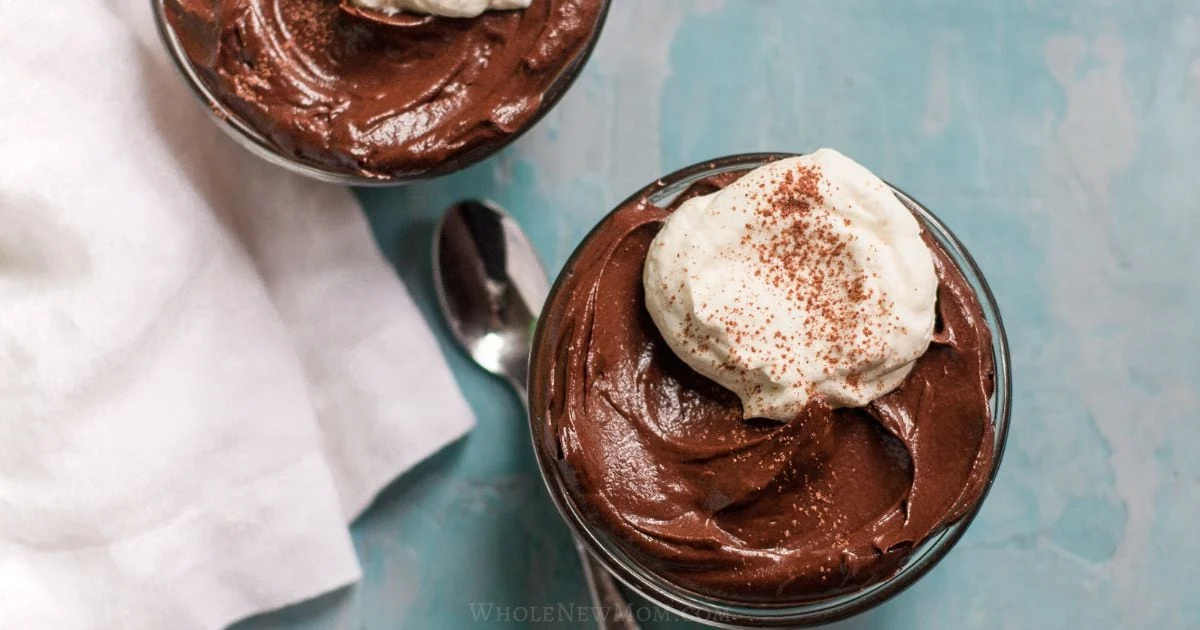 Two glass bowls of chocolate mousse topped with whipped cream and a sprinkle of cocoa powder, next to a spoon and white cloth on a blue surface.