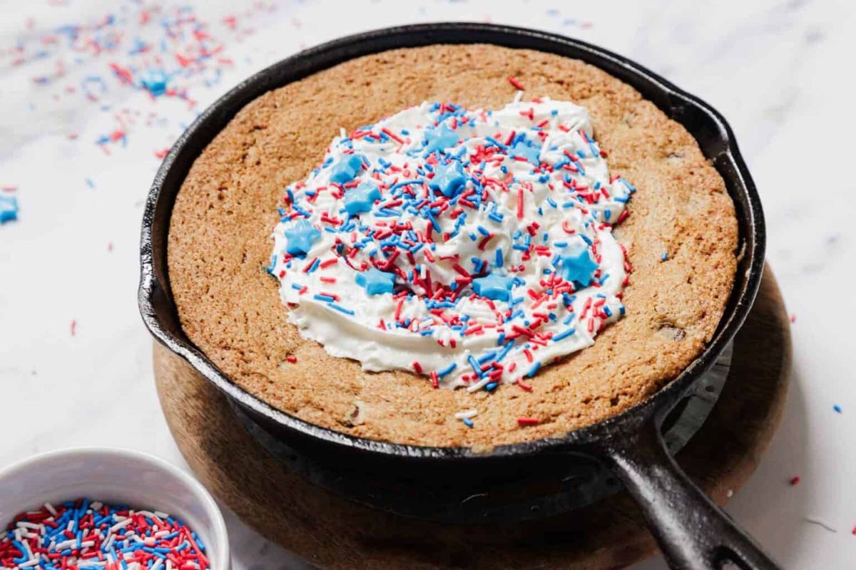 A large cookie in a cast iron skillet, topped with whipped cream and red, white, and blue sprinkles on a wooden board.