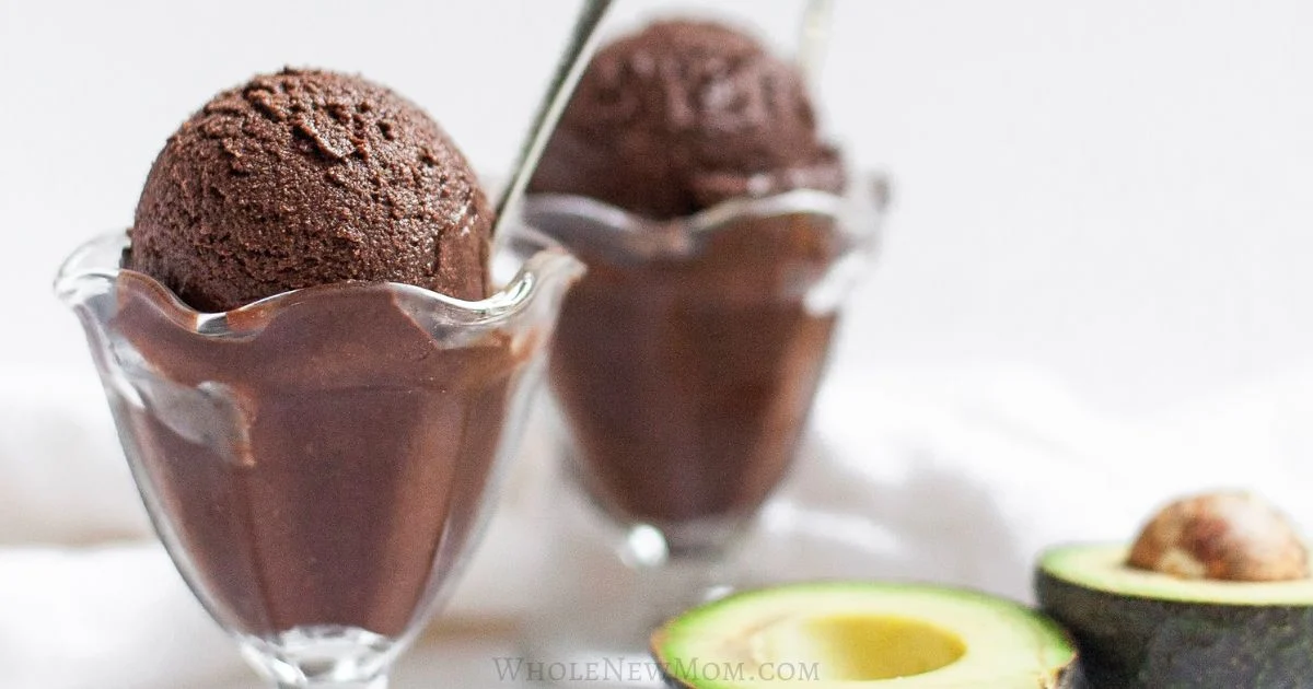 Two glass bowls filled with scoops of dark chocolate ice cream, each with a spoon, next to a halved avocado on a white surface.