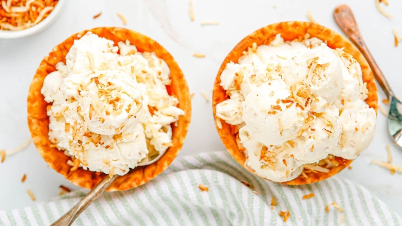Two bowls of vanilla ice cream topped with toasted coconut flakes, each with a spoon, placed on a white surface with a striped cloth nearby.