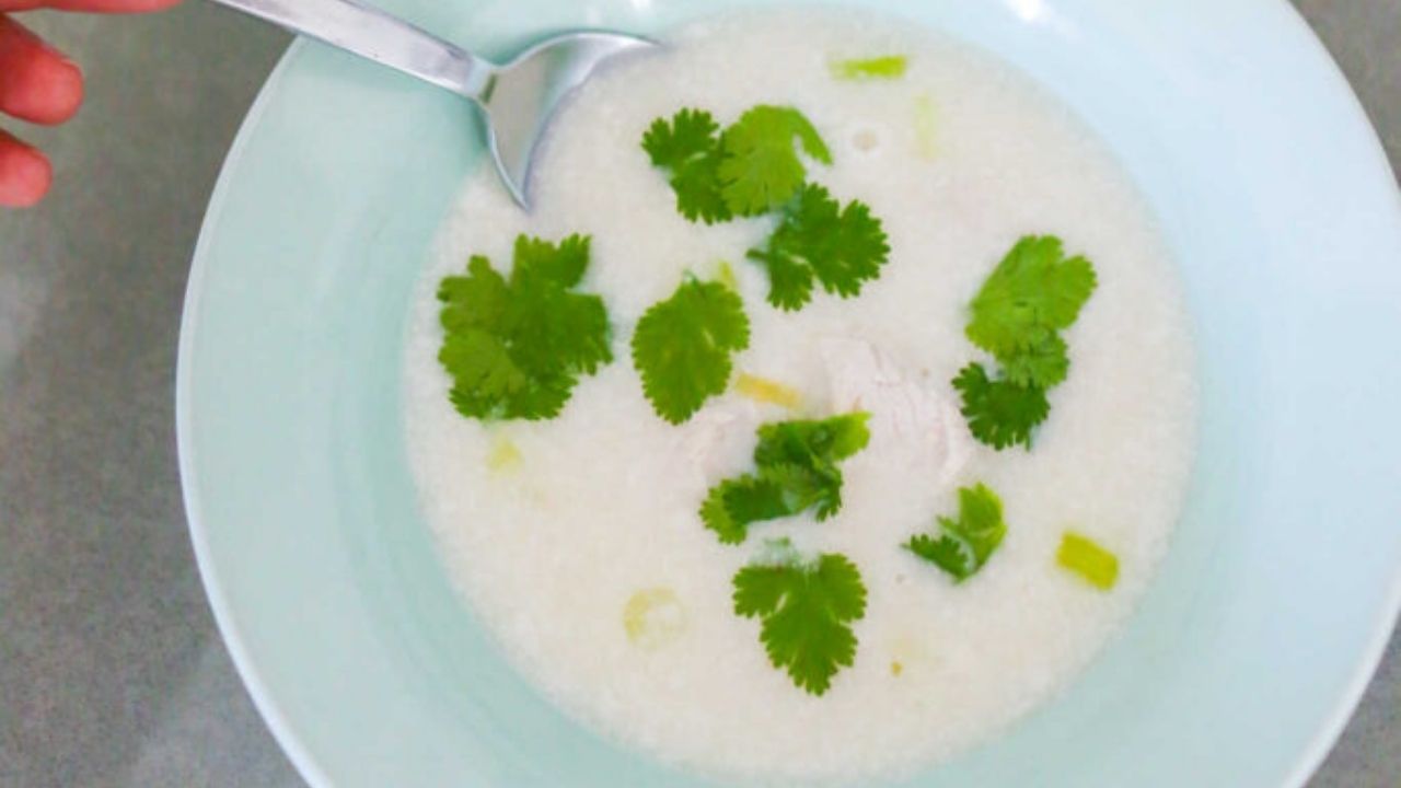 A bowl of rice porridge garnished with cilantro leaves, with a spoon and hand visible on the side.