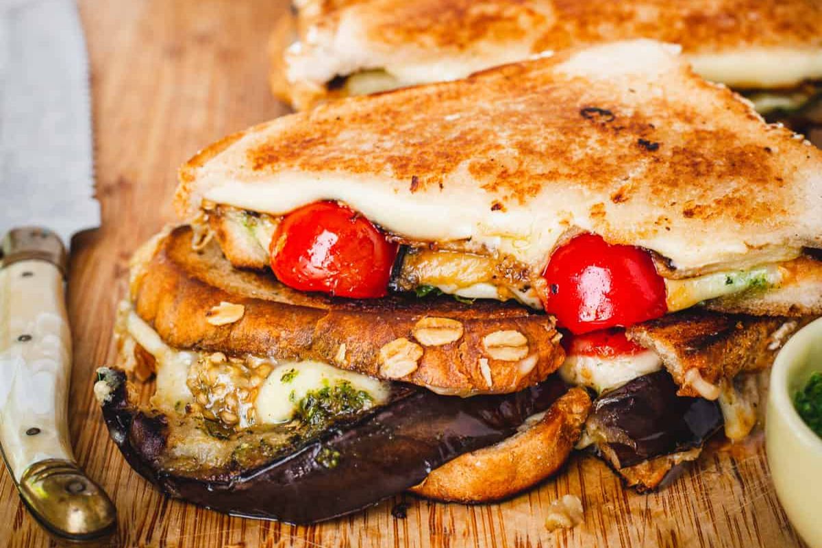 Grilled sandwiches with melted cheese, roasted eggplant, tomatoes, and pesto on toasted bread, placed on a wooden cutting board beside a serrated knife.