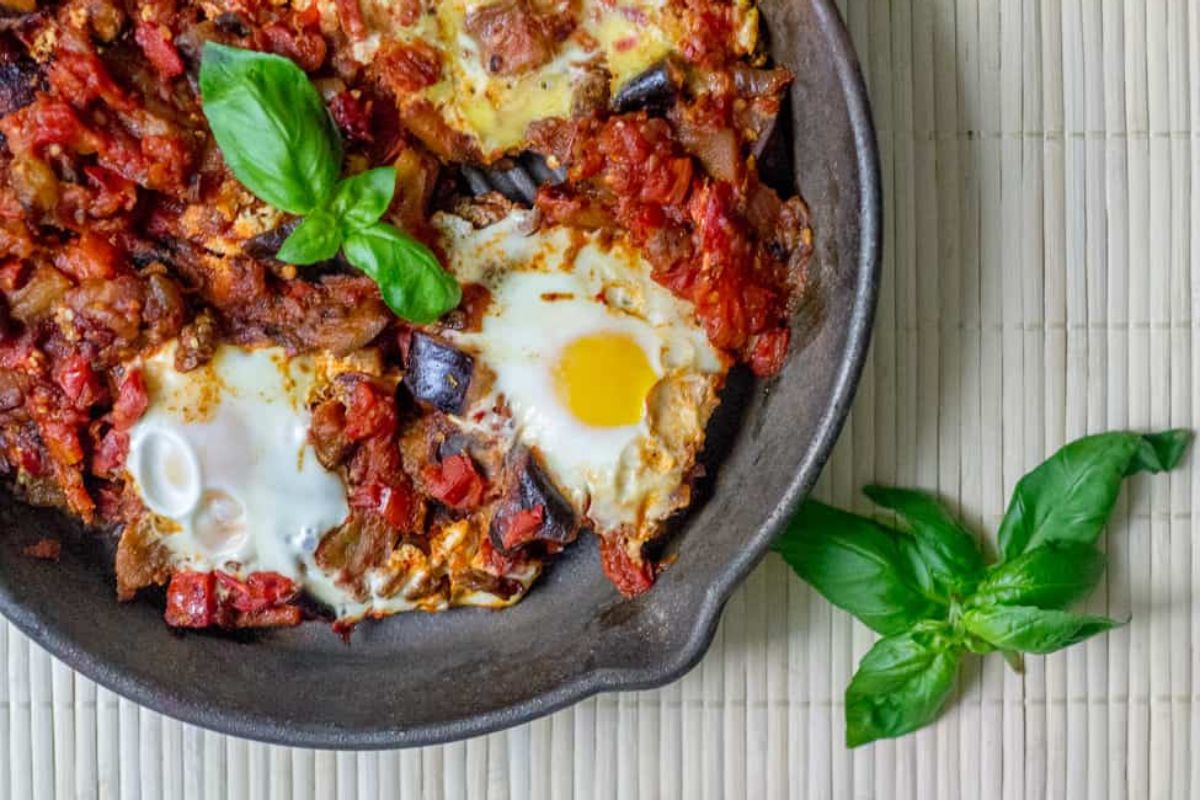 A skillet with baked eggs, tomatoes, and eggplant, garnished with fresh basil, sits on a light bamboo mat with a sprig of basil beside it.