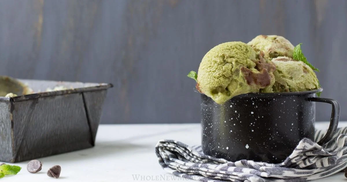 A black speckled mug filled with scoops of green and brown ice cream sits on a striped cloth next to a metal loaf pan.