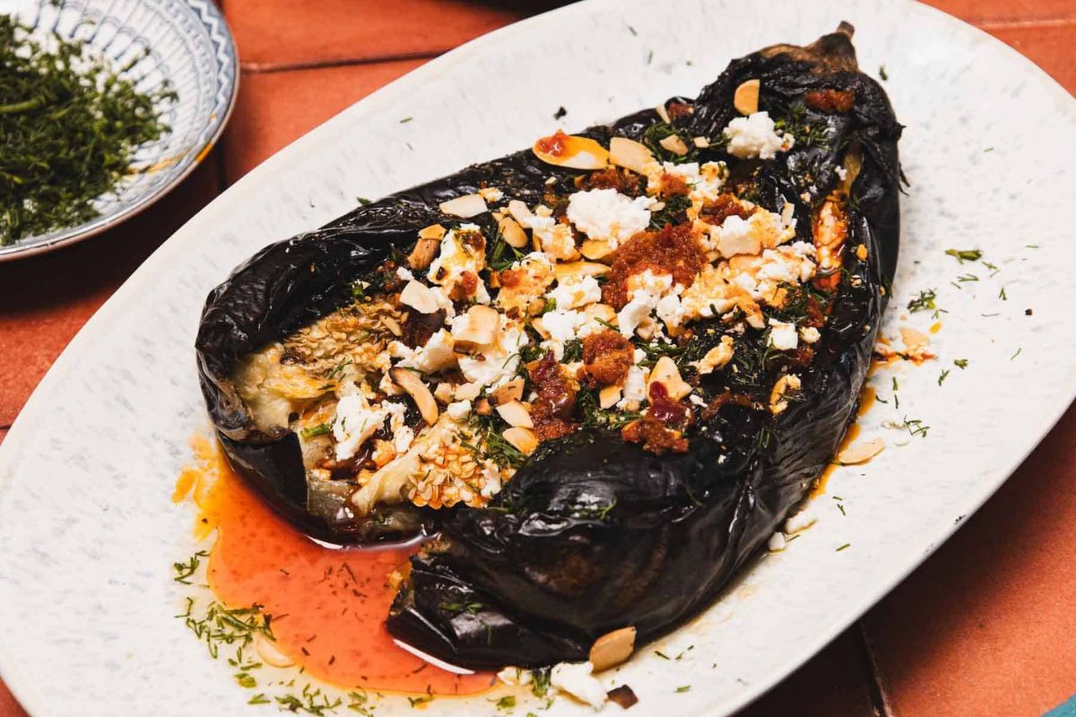 A roasted eggplant topped with crumbled cheese, chopped nuts, herbs, and a drizzle of oil, served on a white oval plate.