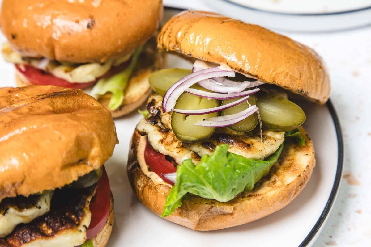 Two grilled veggie burgers on buns with lettuce, tomato, pickles, and sliced red onion, served on a white plate.