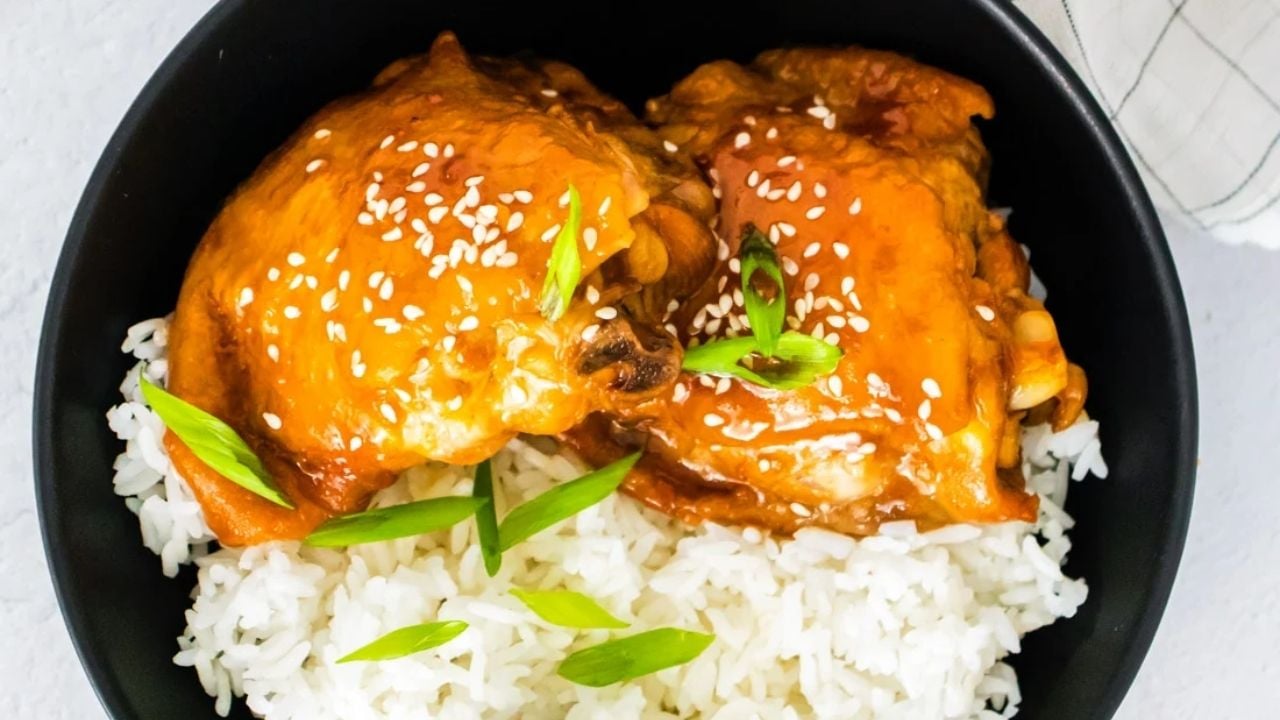 A black bowl filled with white rice topped with two pieces of glazed chicken, garnished with sesame seeds and chopped green onions.